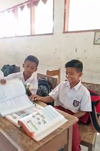 The video shows a classroom environment with several students. The students are wearing school uniforms, consisting of white shirts and red bottoms. In the foreground, two boys are seated at a wooden desk, engaged with open books. One boy on the left is holding a small object and looking at his book. The boy on the right is smiling and looking towards his book. Behind them, other students are visible, also seated at desks and appearing to be studying or working on assignments. Some students are wearing hijabs. The classroom has wooden desks and chairs, and the walls are a light color. There are windows with wooden frames along one side of the room, allowing natural light to enter. A bulletin board with colorful decorations is visible on the wall in the background. The overall scene depicts a typical classroom setting during a lesson or activity.