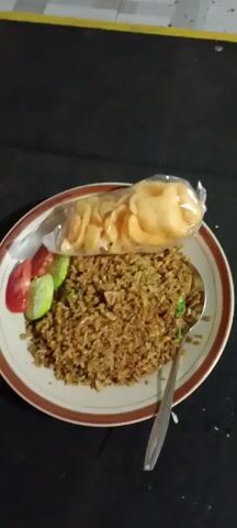 A plate of fried rice, garnished with tomato and cucumber slices, sits on a dark surface. A bag of crackers is placed on top of the fried rice. A spoon is resting on the plate. A glass of dark liquid, presumably tea, is positioned to the left of the plate. A box of tissues is visible to the left of the glass. The video appears to be shot in a dimly lit environment. The timestamps indicate that the scene is being recorded for a short duration, with a slight camera movement suggesting a slight pan or tilt.