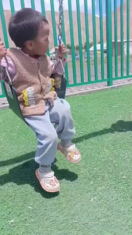 A young child with dark hair is sitting on a green swing, holding onto the chains with both hands. The child is wearing a pink sweater, a brown vest with a cartoon image, light blue pants, and white and pink shoes with cartoon characters on them. The swing is set against a background of a green fence, a grassy area, and a mountainous landscape under a blue sky with some clouds. The child is looking to the right.
