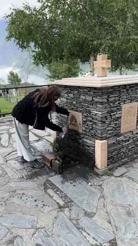 A person wearing a black knitted sweater and light-colored pants is filling a clear plastic water bottle from a brass spigot attached to a stone structure. The spigot is ornate and appears to be a fountain. The stone structure has a layered, rough texture and a decorative carving on its right side. Above the spigot, there is a circular relief carving. Atop the structure, there is a light-colored cross. The ground is paved with irregular stones, and water is flowing from the spigot into the bottle and splashing onto the stones below. A pile of bricks is near the base of the fountain. In the background, there are trees with green foliage and a hazy, mountainous landscape. The lighting suggests an overcast day.