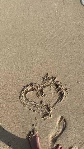A close-up, overhead shot shows a heart drawn in the sand on a beach. The tide is coming in, and a wave is about to wash over the heart. A person's bare feet are visible at the bottom of the frame.