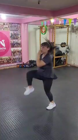 A woman with dark hair tied in a ponytail, wearing a dark gray t-shirt and dark gray leggings with white sneakers, is performing a series of energetic dance or exercise moves in a gym. The gym has a black rubber floor and pink walls. In the background, there are posters on the wall, a weight rack with weights and kettlebells, and colorful flags strung across the ceiling. The woman is in motion throughout the video, kicking, jumping, and spinning.