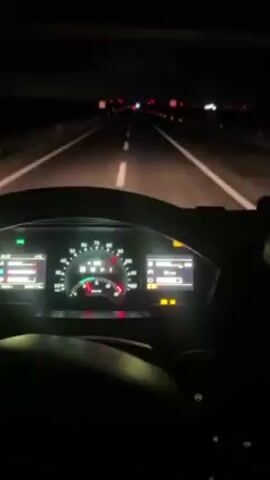 The scene captured from within a vehicle, shows the view through the windshield driving at night on a multi-lane highway. The dashboard instruments glow in the dark cabin, providing information about the vehicle's speed and other indicators. Out of the windshield, the road stretches forward, marked with white lines. Headlights from the vehicle illuminate the road ahead, and red tail lights of distant vehicles are visible in the dark background. The environment is dark, with minimal ambient light except for the streetlights and the tail lights of other cars.
