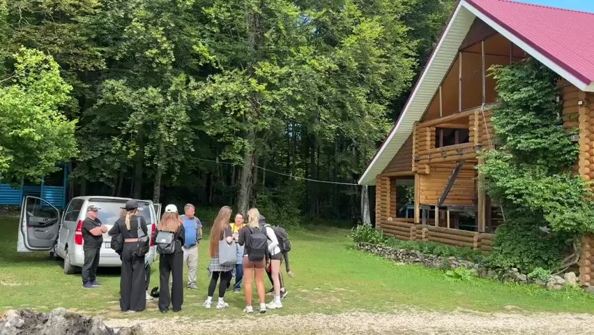 A group of people are gathered outdoors near a wooden house and a minivan. The house is made of logs with a red roof and is surrounded by greenery. A silver minivan with its doors open is parked to the left of the house. Several individuals, including both men and women, are standing around, some holding backpacks. One man appears to be speaking to a group of younger people, possibly giving instructions or information. The environment is natural, with trees and grass visible.