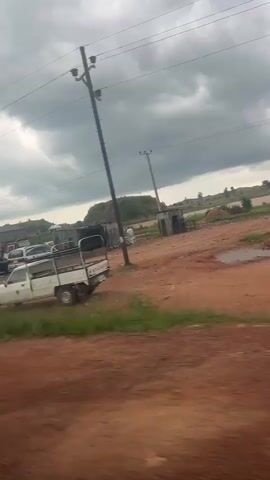 The video pans across a dusty, outdoor scene, likely a construction or storage area. Several vehicles are visible, including a white pickup truck with a flatbed loaded with what appears to be building materials, a white van, a dark colored sedan, and a red car. There are piles of dirt or sand and stacks of what look like bricks or concrete blocks. Several simple, unfinished structures are present, some with corrugated metal roofs. Overhead power lines are visible, and the sky is cloudy. In the background, there are some trees and what looks like a large mound of earth or debris. The ground is red, dusty earth. A person in a light-colored shirt is visible near one of the structures. A three-wheeled truck is also present.
