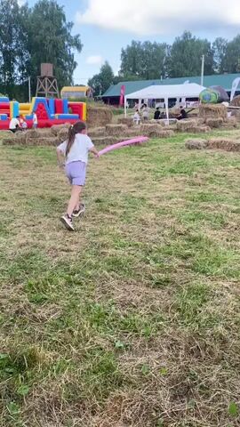 A girl in a white t-shirt and purple shorts runs across a grassy field holding a long pink balloon. In the background, several children are interacting with costumed characters. One character is dressed as a unicorn and another as a yellow rabbit. The children are holding long balloons, possibly used as swords, and appear to be engaged in a playful sword fight. The environment includes hay bales scattered around the field and some inflatable structures in the distance. The sky is partly cloudy.