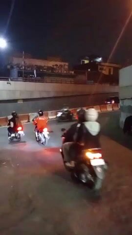 This is a nighttime video showing several motorcycles on a paved road. The motorcycles are moving in the same direction, and some of them have their brake lights illuminated. The road appears to be in an urban area, with buildings and a bridge visible in the background. The scene is dimly lit, with streetlights providing some illumination.