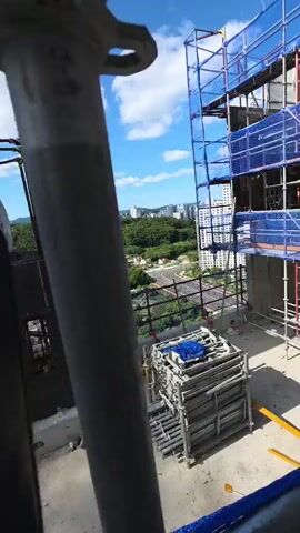 This media file captures a construction site on a bright, sunny day with a blue sky and scattered white clouds. The scene is set high up, offering a panoramic view of a lush green landscape dotted with buildings and a distant cityscape. In the foreground, scaffolding structures, draped with blue netting, are prominent, indicating ongoing construction on a tall building. Various construction materials, including metal pipes, wooden planks, and a cart filled with metal components, are scattered across the concrete surface. A person, dressed in blue, can be seen sitting on a ledge on the left side of the frame, partially obscured by a concrete structure. The overall atmosphere is one of active development under favorable weather conditions.