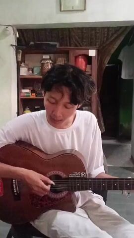 A young man with dark, messy hair is sitting and playing an acoustic guitar. He is wearing a white t-shirt and light-colored pants. The guitar is brown with a red pickguard and some stickers on it. He is strumming the guitar with his right hand and holding the neck with his left. The background appears to be an indoor space, possibly a room, with a bookshelf behind him filled with various items. There is also a curtain visible. The lighting suggests it is daytime.