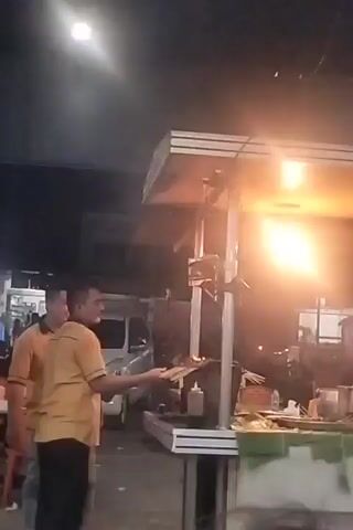 A short video displays a nighttime scene with a food stall. A man in an orange shirt is shown holding skewers and what appears to be a plate, standing next to a grill where food is being cooked. The grill has a bright light source, possibly from the cooking flames or overhead lighting, which illuminates the area. Another person in a similar orange shirt is visible in the background. The stall is set up outdoors, with some vehicles and other structures present in the dimly lit surroundings. The overall atmosphere suggests a busy street food vendor operation.