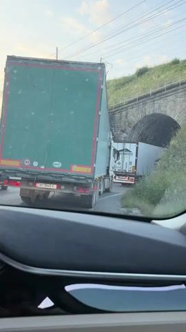 The video shows a view from inside a car looking out at a road. There are several large trucks on the road, with the closest truck's green trailer filling the majority of the frame. The truck appears to be stationary or moving very slowly. In the background, a tunnel entrance is visible, with another white truck entering it. Other vehicles and roadside vegetation are also present. The road curves to the right, leading into the tunnel. The time stamp indicates the video is from June 11, 2023, around midnight.