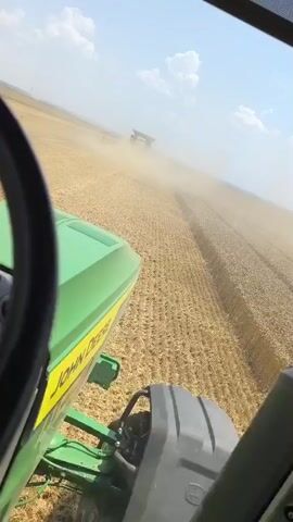 The video shows a first-person perspective from the cab of a green tractor, labeled "JOHN DEERE", moving through a field of ripe wheat. Dust is kicked up by the tractor and a red combine harvester operating ahead and to the right. The combine is actively harvesting the wheat. A digital display on the tractor shows numerical data, and a tablet-like screen is also visible. The sky is blue with scattered clouds. The environment appears to be a large agricultural field on a sunny day.