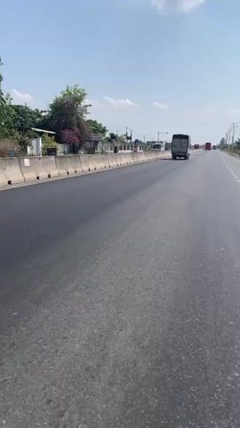 The video depicts a wide, straight, asphalt road under a clear blue sky with a few scattered white clouds. Along the left side of the road, there is a low concrete barrier, behind which are some buildings, greenery, and power poles. Several trucks are visible in the distance, driving in the same direction as the camera. The asphalt is mostly black, with some lighter gray patches and a faded white lane marking. There is no indication of the camera's exact location, but the setting appears to be a rural or semi-rural area.