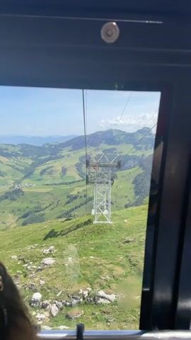 The video shows a view from inside a cable car as it passes a support tower. Outside the cable car, a green mountainous landscape stretches into the distance under a bright blue sky with scattered clouds. The support tower is made of a white metal structure with a red spherical object on top. The interior of the cable car is partially visible with a person's head in the lower left corner and a hat appearing later in the lower right corner. The camera remains stable and focused on the view outside the window.