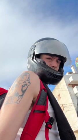 The video shows a person wearing a black helmet with a clear visor, a white tank top, and a red backpack with black straps. The person has tattoos on their left arm, including one with the name "NANCY" visible. They also have piercings on their nose and small tattoos on their face. They are on a motorcycle, and part of another motorcycle and rider is visible to the right. The background shows a blue sky with some clouds and a few buildings. The person is looking directly at the camera at the beginning of the video, then turns their head to the right.