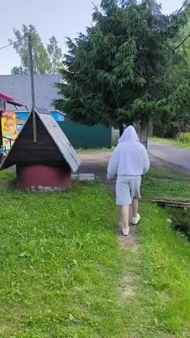 A person wearing a white hoodie and grey shorts walks away from the camera on a dirt path. The path is lined with green grass and a large evergreen tree. To the left of the path is a wooden well with a red base and a metal roof. Behind the well is a small wooden building with a red roof and a sign that reads "ПРОДУКТЫ" in red letters. A wooden bench is in front of the building. In the background, there are houses and a green fence. The sky is clear and blue.