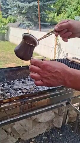A close-up shot shows a person's hands pouring dark liquid from a copper pot with a long, ornate handle into a small, clear glass. The pot is tilted, and the liquid streams into the glass, which is held steady by another hand. The scene appears to be outdoors, with a charcoal grill in the foreground. The grill contains hot embers, and the setup suggests the coffee may have been prepared or is being kept warm over the heat. In the background, there are trees and a stone wall, indicating a backyard or outdoor setting. The lighting suggests it is daytime.