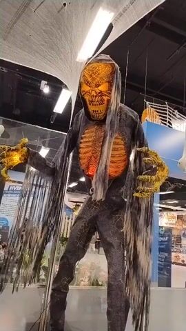 A tall, animatronic Halloween decoration stands center frame. It depicts a skeletal figure with a hooded robe, tattered sleeves, and a grinning, skeletal face with glowing orange eyes. Its bony hands are outstretched and also glow orange. The figure's chest shows a ribcage with a pulsating red light in the center. The background appears to be an indoor store, possibly a Halloween or party supply store, with ceiling lights, displays, and other store merchandise visible. White, wispy decorations that resemble spiderwebs hang from the ceiling above the figure.