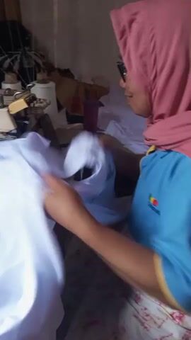 A woman wearing a pink hijab and blue shirt is seen sewing a white garment on a sewing machine. She is seated at a table with various sewing supplies in the background. The woman's hands are holding the fabric as she guides it through the sewing machine. The video is a close-up shot, focusing on the sewing process. The environment appears to be a home or small workshop setting.