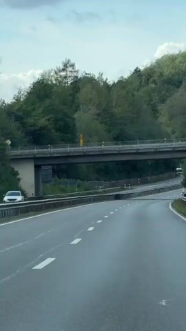 The video shows a highway underpass with a bridge overhead. The highway has multiple lanes and is surrounded by trees and greenery. There are white lines marking the lanes and guardrails along the sides of the road. A few cars are visible on the highway, and there is a pedestrian bridge above the road. The sky is overcast.