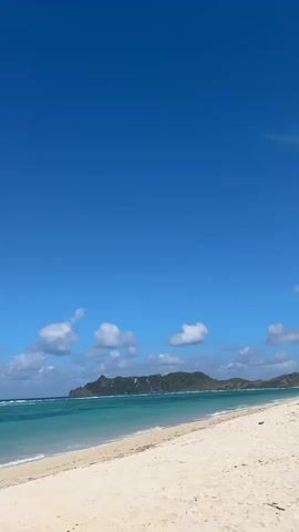 The video shows a serene beach scene under a clear blue sky. The turquoise ocean meets a white sand beach, with a distant green island visible on the horizon. Gentle waves lap at the shore. A man wearing a dark baseball cap and sunglasses is positioned in the foreground, facing away from the camera. He is holding a smartphone in both hands, appearing to take a photograph or video of the ocean. The man is shirtless, and a watch is visible on his left wrist. The camera perspective is from behind and slightly to the side of the man, focusing on the beach and the ocean beyond.