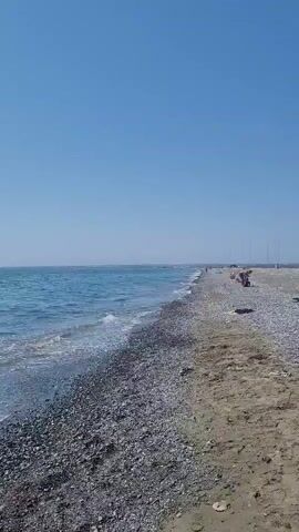 The video shows a wide, unobstructed view of a beach on a clear, sunny day. The ocean on the left is a vibrant blue with gentle waves lapping at the shore. The beach itself is a mix of sand and pebbles, with a distinct line separating the two textures. In the distance, along the pebble section of the beach, several figures can be seen. One person is sitting upright, facing away from the camera, while another appears to be lying down further down the beach. The horizon is a clear, pale line under a vast expanse of bright blue sky. There are no discernible structures or objects other than the natural elements of the beach and the distant figures. The camera remains stationary, offering a static panoramic view.