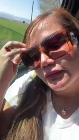 A woman with long brown hair is in a car wearing large brown-tinted sunglasses and a white shirt with a gold necklace. Her lips are colored with a deep red matte lipstick. She is holding the glasses with her left hand, which has a red gemstone ring. The background shows a green landscape under a bright blue sky. The camera zooms in on her face as she puckers her lips.