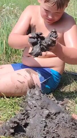 This media file features a young boy, likely of European descent, playing in mud outdoors. He is shirtless and wearing blue swimming shorts. The boy is seated on the ground, which is covered in grass. He is actively engaging with a pile of mud, scooping it with his hands and smearing it onto his chest and legs. His face shows concentration as he plays. The background consists of lush green vegetation, suggesting a rural or natural setting, possibly during warmer months due to the greenery. The lighting indicates it is daytime, with bright sunlight casting shadows, hinting at a clear day. The overall scene conveys a sense of childhood innocence and engagement with nature. There is no visible text in the media file. The boy appears to be enjoying himself, as indicated by his focused yet relaxed posture.