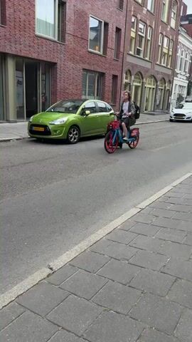 A person is riding a blue and red bicycle with a basket on the front and a suitcase attached to the back, down a cobblestone street. The person is wearing a green jacket and shorts, with a backpack on. They are looking to their right. Buildings with brick facades line the street. A green car is parked on the left side of the street.