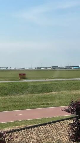 The video shows an airport scene with airplanes parked on a tarmac in the distance. In the foreground, there is a grassy area with a red running track. A chain-link fence runs along the bottom of the frame, partially obscuring the view. A red and yellow sign that reads "STOP" is visible in the middle ground. The sky is blue with some scattered clouds. The weather appears to be sunny and clear.