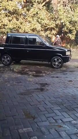 A man in a cap and shirt stands next to a black four-door pickup truck. The truck is parked on a dark brick surface. There are trees with green and yellow leaves in the background. The man appears to be cleaning or inspecting the truck.