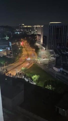 The video shows a nighttime cityscape viewed from an elevated perspective. The scene is dominated by streets with traffic lights and moving cars. A wide, divided road runs through the frame, curving slightly. Cars with headlights on are visible, moving along the road. Adjacent to the road is a green space with trees and some illuminated concrete structures. In the background, multiple tall buildings are visible, lit up against the night sky. The camera remains static throughout the video.