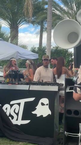 The video shows an outdoor party scene. A DJ stands behind a black DJ booth with a white "OST" logo and a ghost graphic. He wears a light beige shirt, a cap with a "Monster Energy" logo, and has tattoos on his arms. He takes a sip from a yellow drink. Several people are standing around the DJ booth, including two women with headbands and drinks, a woman in a white corset dress with tattoos on her back, and other party-goers. A large, white, horn-shaped speaker stands to the right of the booth. Palm trees are visible in the background, and a white tent offers shade to some of the attendees. The overall atmosphere is festive and lively.