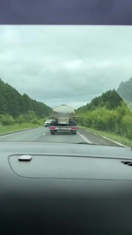 A video taken from the interior of a car shows a large tanker truck driving on a two-lane road surrounded by trees. The truck is silver and has yellow reflective tape around its tank. The car is following the truck, and another car is visible in the left lane. The sky is cloudy. The license plate on the truck is visible and reads "AP 9521 18".