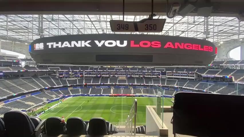 The video shows an empty soccer stadium with a large digital scoreboard displaying the message THANK YOU LOS ANGELES in white and red letters. The stadium seats are dark blue and appear unoccupied, except for a few individuals in the background on the field. The field itself is green and marked for soccer. Above the scoreboard, two small digital displays show numbers 349 and 350. The stadium has a large, open roof structure. The perspective is from a seating area looking down towards the field.