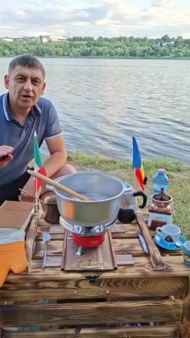 A man is sitting at a wooden table by a lake and is looking at the camera and talking. On the table are a pot of water on a stove, a coffee maker, a coffee grinder, a cup and saucer, and a bottle of water. There are also two flags, one Italian and one Romanian, and a book. The man is wearing a dark blue polo shirt and black shorts. The lake is calm and the sky is overcast. The man is gesturing with his hands as he talks.