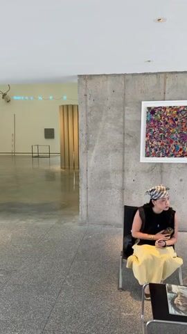 The video opens with a view of an art gallery or museum. On the right, a woman wearing a black sleeveless top, a yellow skirt, and a patterned headscarf is seated on a chair. She is looking down and holding something in her hands. Her bag is slung over her shoulder. The background shows a large, open space with a polished concrete floor. There are several art installations visible: a large gold cylindrical sculpture in the center, a framed artwork on the wall to the right of the sculpture, and a smaller square artwork on the wall to the left. There is also a neon sign with blue lights in the upper left corner, and what appears to be a deer head mounted on the wall. Natural light streams in from an opening at the far end of the gallery, revealing greenery outside. The camera pans slowly across the scene, offering a wider perspective of the gallery space.