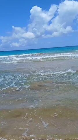 The video presents a tranquil beach scene on a partly cloudy day. Gentle waves lap against the sandy shore, and a large, modern hotel building stands in the background to the right.  The beach appears mostly deserted.  A person’s voice states "Luquillo" .