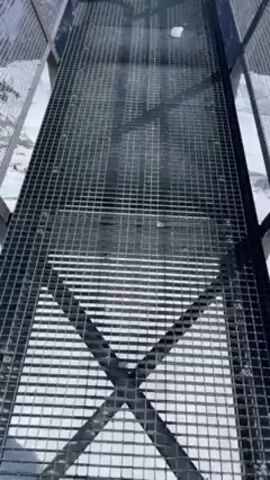 The video shows a person walking on a metal grid platform towards a viewpoint overlooking a snowy mountain range. The camera pans up to reveal a sign that says "La Clusaz". The sky is clear and blue. The mountains are covered in snow and are visible in the distance. The platform has metal railings on either side. There is snow visible beneath the metal grid platform.