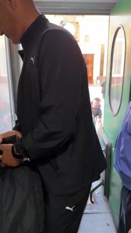 A man steps into a train car carrying a box labeled "EMERALD" and then turns, walks into the car, and passes by a man wearing a blue shirt. The man carrying the box has a black bag slung over his shoulder. The background shows a train station.