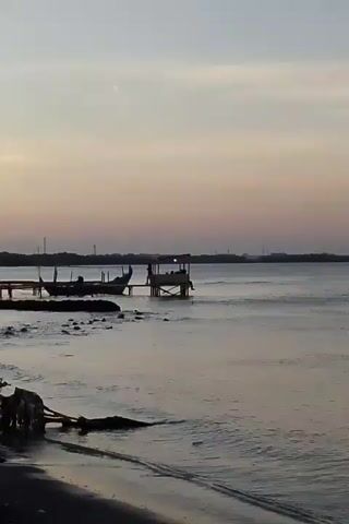 The video shows a serene coastal scene at dusk. The sky is a soft gradient of pink and grey, with a hint of a moon visible. The water is calm, with gentle ripples lapping at a dark sand beach in the foreground. On the beach, there appears to be some debris, possibly driftwood and trash. In the middle ground, a wooden pier extends into the water, with two boats moored alongside. Further out on the pier, a small structure with a thatched roof serves as a shelter, and a few people are gathered there, silhouetted against the dim light. The horizon line is indistinct, with a low-lying landmass visible in the distance. The overall atmosphere is peaceful and tranquil.