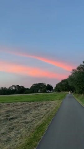 A gentle breeze rustles the leaves of trees lining a paved path as two people cycle into the distance. The path winds through a rural landscape, bordered by lush green fields and a stretch of harvested hay. The sky above is a canvas of soft pastels, transitioning from a pale blue at the zenith to hues of pink and orange near the horizon. Scattered clouds catch the last rays of the setting sun, painting them with fiery colors. In the background, a cluster of houses with red-tiled roofs nestles amidst the rolling hills, suggesting a tranquil village setting. The overall atmosphere is one of peacefulness and natural beauty, capturing a serene moment at dusk in Bissendorf, Germany.