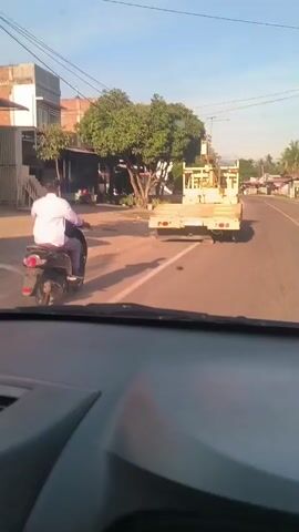 The video shows a view from inside a car driving down a street. A motorcycle passes the car on the left. Ahead, a large yellow vehicle with a "TADANO" crane is driving slowly in the same direction. The street is lined with buildings, trees, and utility poles. The sky is bright and clear.
