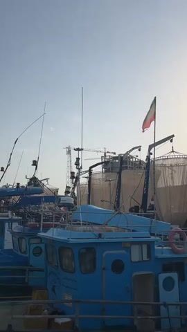 The video displays a row of blue fishing boats docked in a harbor. The sky is clear and blue with a few wispy clouds. An Iranian flag is visible on one of the boats. In the background, industrial structures, cranes, and a large arched bridge are present. The water is calm and reflects the sky and the boats. The focus is on the boats and the surrounding port environment.