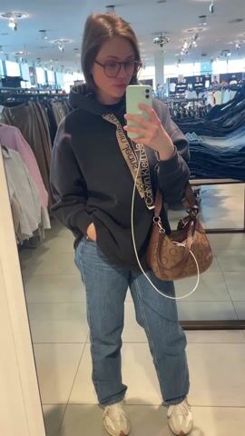 A woman is taking a mirror selfie in a clothing store. She is wearing glasses, a dark grey hooded sweatshirt with "Calvin Klein Jeans" printed on the hood, blue jeans, and beige sneakers. She has a brown purse with a light brown strap that also has "Calvin Klein" printed on it, and a white phone charger cord is hanging from the bag. She is holding a light green phone in her left hand. In the background, there are racks of clothes and shelves of jeans. The floor is tiled. The lighting is bright.