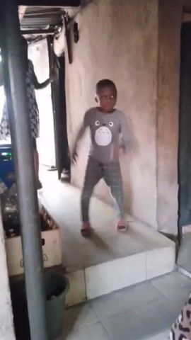 A small child is dancing on a tiled floor in what appears to be a home. The child is wearing a long-sleeved grey shirt with a cartoon bear on it and grey plaid pants. They are also wearing pink sandals. The child dances expressively, lifting their arms and legs in time to music that can be heard. Behind them, partially obscured by a wooden post, a taller person wearing a patterned dress is also dancing. The room has a low ceiling, and the walls are unfinished. There are a few items in the background, including a crate and a green bucket.
