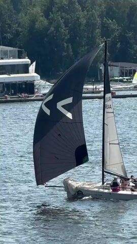 The video shows a sailboat on a body of water. The sailboat has two sails, one large black sail with white markings and a smaller white sail. There are several people on the sailboat. In the background, there are other sailboats, a building, trees, and a dock. The water is rippling, and the sky is visible. The scene appears to be a sunny day.