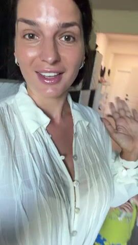 The video features a woman with dark hair and light skin, wearing a white, open-collared blouse. She is indoors, likely in a kitchen or living area, with cabinetry and a doorway visible in the background. The woman is smiling and speaking directly to the camera, waving at one point. She mentions living in Miami with her family. The lighting is bright, and the overall atmosphere appears casual and personal.