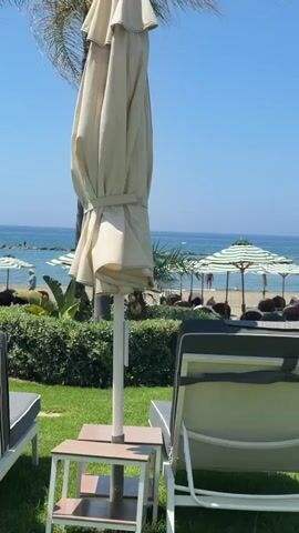 The video shows a beach scene with rows of lounge chairs and umbrellas on a grassy area. The ocean and the sky are visible in the background. Some people are sitting on the lounge chairs, while others are walking on the beach. The overall atmosphere is relaxed and vacation-like.