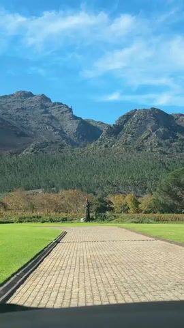 The video shows a stone path leading towards a statue in a garden. Behind the statue is a backdrop of trees and mountains under a blue sky. The statue is of a woman in a long dress and bun, standing on a stone base.  The path is made of beige bricks.  The video is shaky, likely handheld.